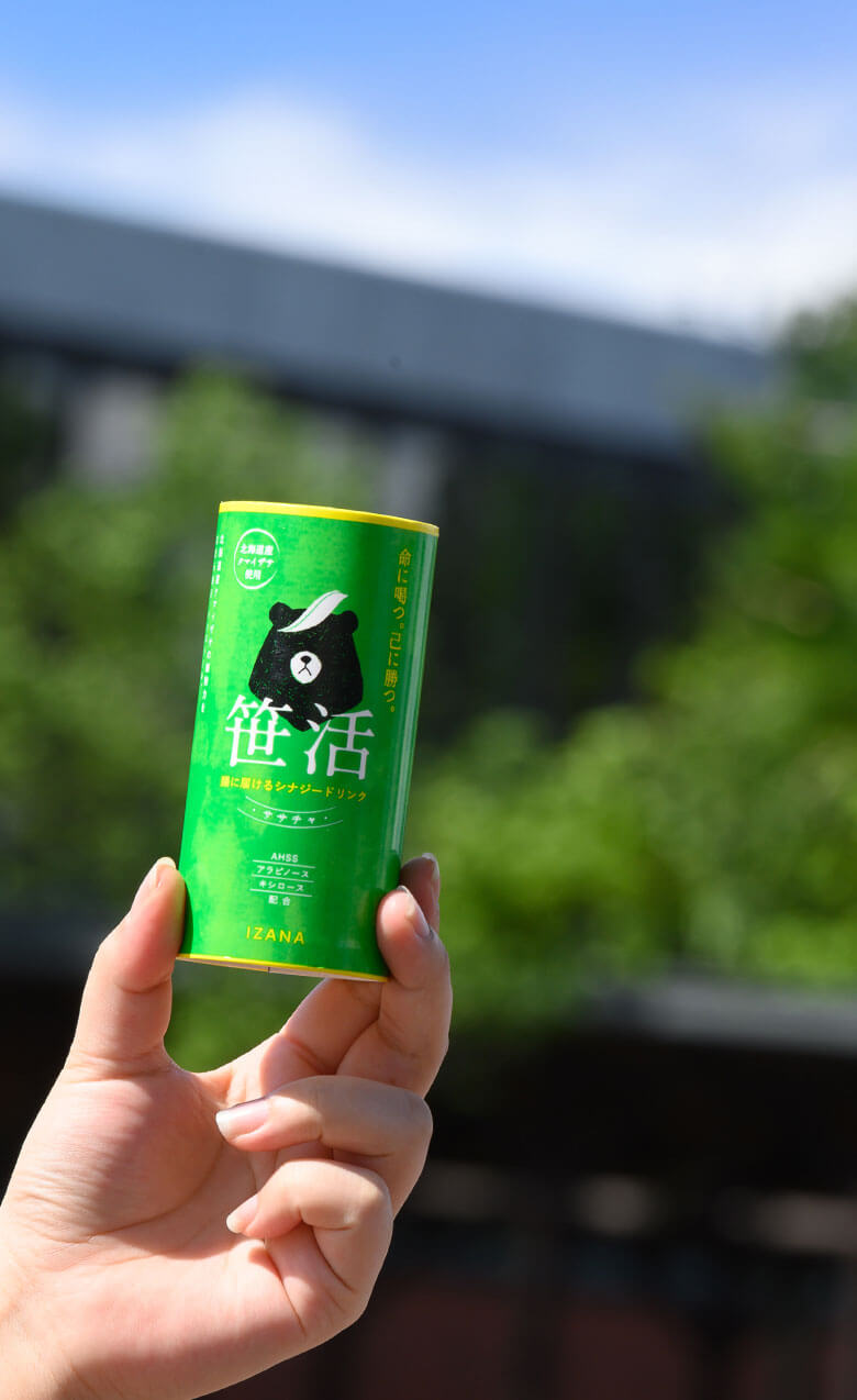 青い空と緑の自然を背景に、笹茶のボトルを持つ人の手が見える。柔らかな自然光が差し込み、明るい雰囲気が漂う。
